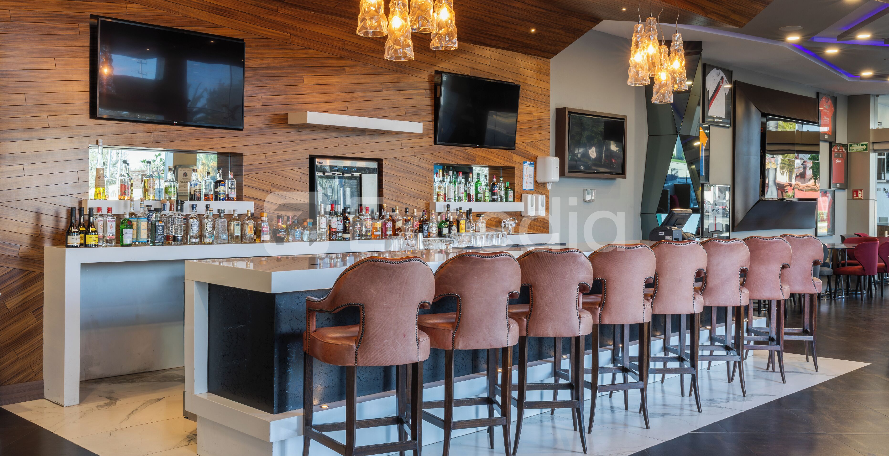 Uma área de bar com um balcão comprido, várias garrafas e banquetas.