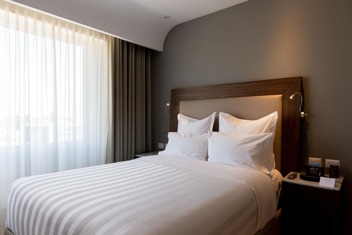 Ein Hotelzimmer mit einem großen Bett, einem Nachttisch mit Telefon und Notizblock sowie einem Fenster mit durchsichtigen Vorhängen.