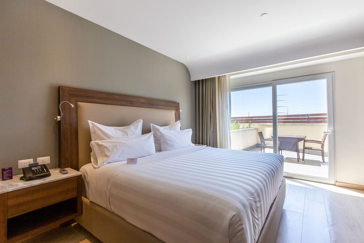 Ein Hotelzimmer mit einem großen Bett, einem Nachttisch mit Telefon und Fernbedienung sowie einem Balkon mit Tisch und Stühlen.