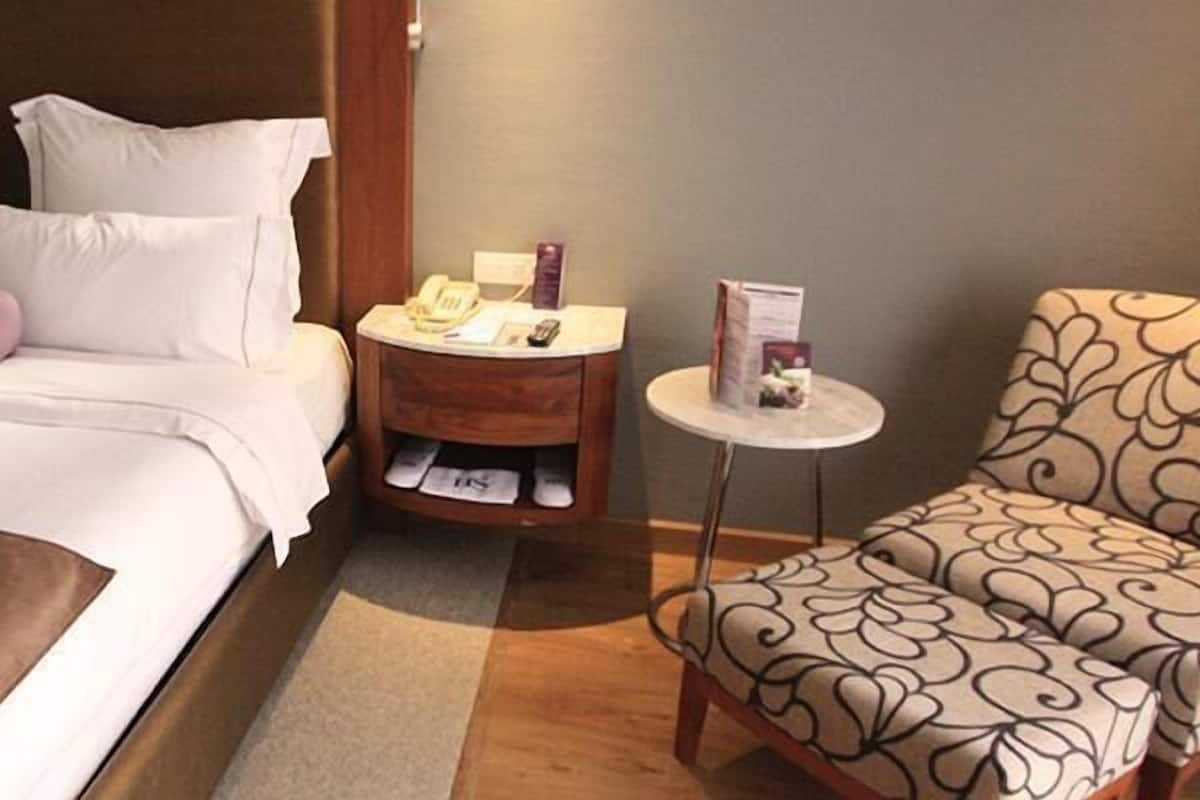 Ein Hotelzimmer mit einem Bett, einem Nachttisch mit Telefon und Zeitschrift, einem Sessel mit gemustertem Kissen sowie einem kleinen Tisch mit einer Lampe.