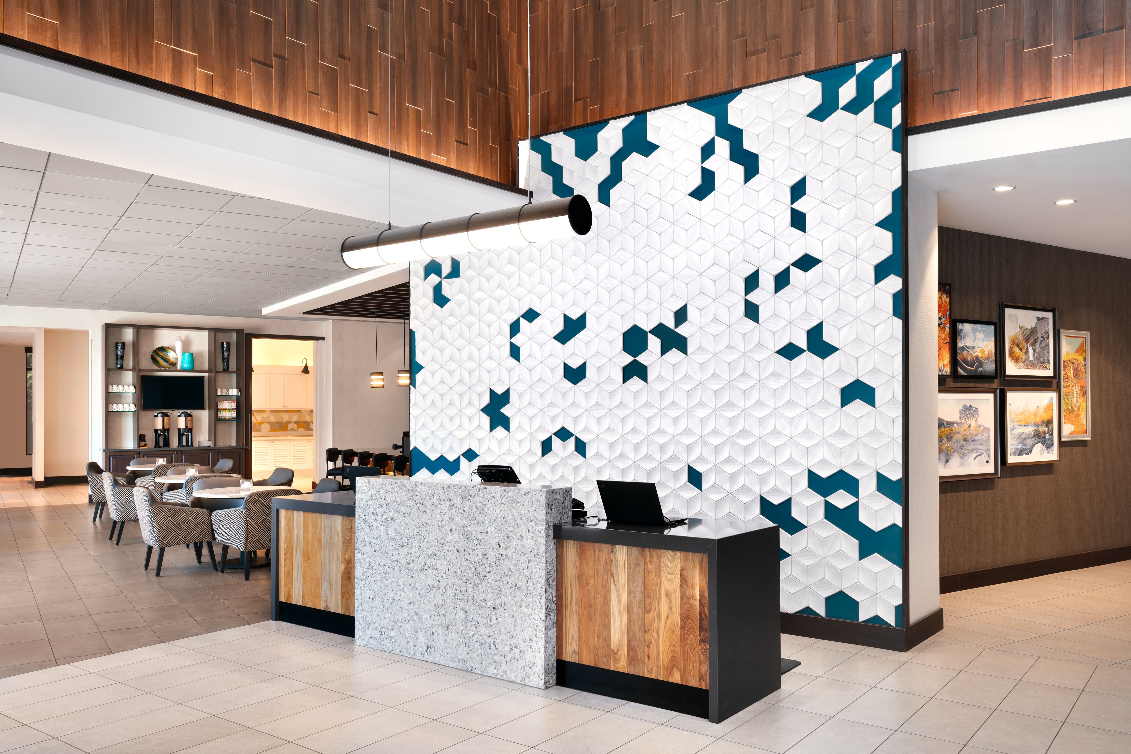 A modern reception desk with a geometric patterned wall, a seating area with chairs, and a visible kitchen in the background.