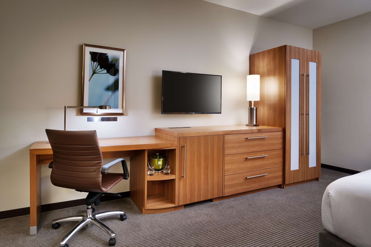 A hotel room with a wooden desk, a flat-screen TV, a lamp, a wardrobe, and a bed.