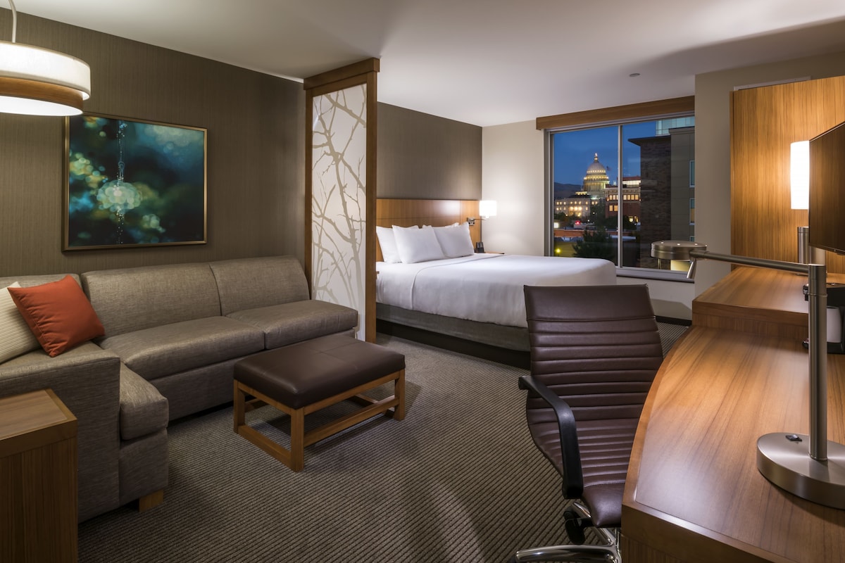 A hotel room with a bed, a sofa, a desk, and a view of a cityscape.