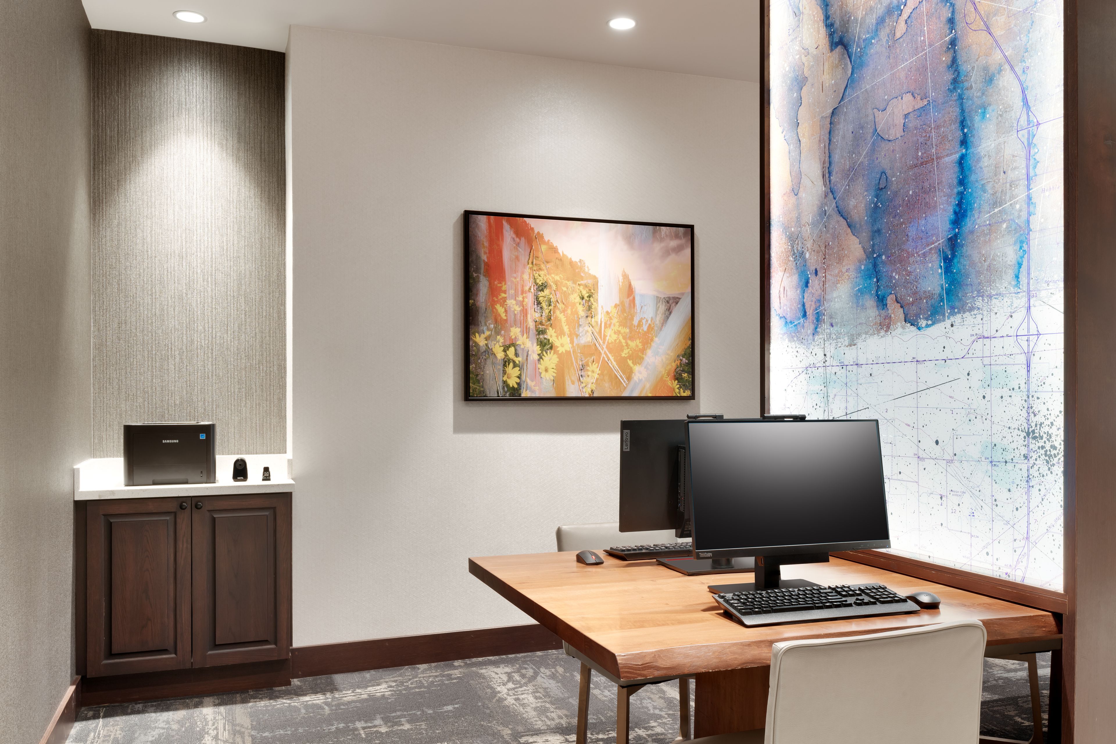 A hotel room with a desk, computer, and artwork on the wall.