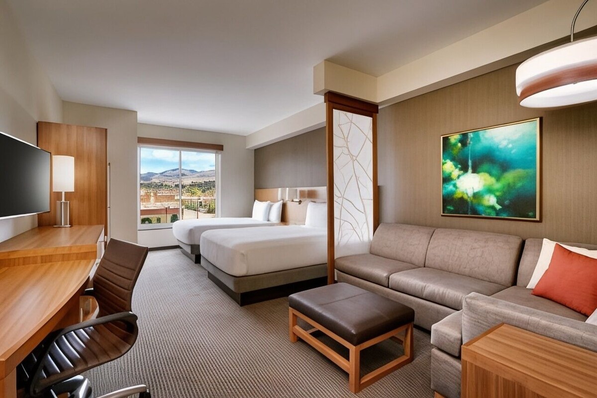 A hotel room with a bed, sofa, desk, TV, and a large window with a view.