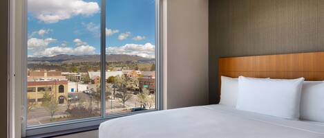Habitación de hotel con un ventanal grande que ofrece vistas a la ciudad, una cama bien tendida y un cabecero de madera.