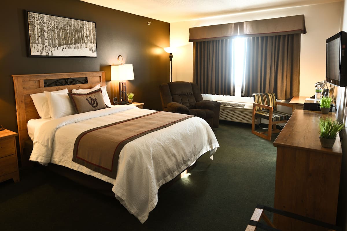 A hotel room with a large bed, two bedside tables with lamps, a desk with a chair, a potted plant, and a large window with curtains.