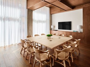 A conference room with a large wooden table, wooden chairs, a TV on the wall, and a large window with sheer curtains.