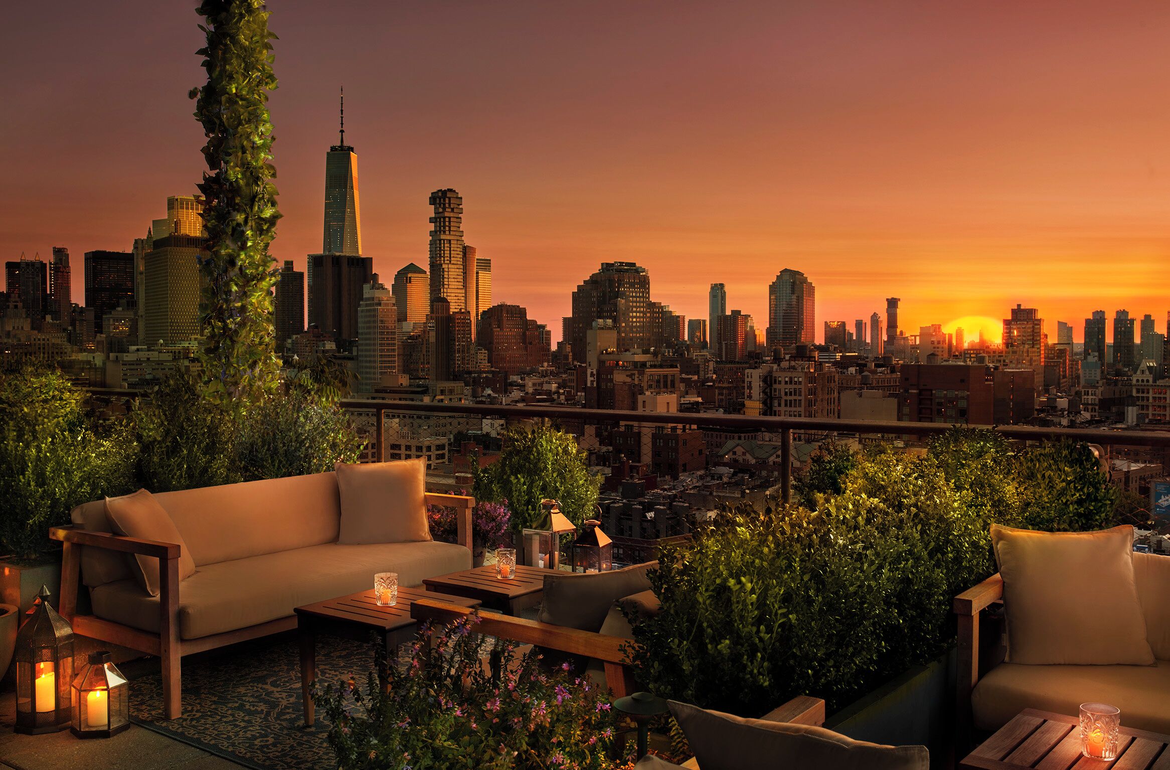 A rooftop terrace with a city skyline view at sunset, featuring a sofa, a coffee table, and potted plants.