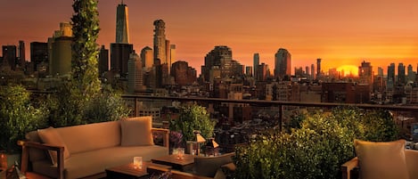 A rooftop terrace with a city skyline view at sunset, featuring a sofa, a coffee table, and potted plants.