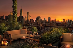 Terraço no telhado com vista para o horizonte da cidade ao pôr do sol, com um sofá, uma mesa de centro e plantas em vasos.