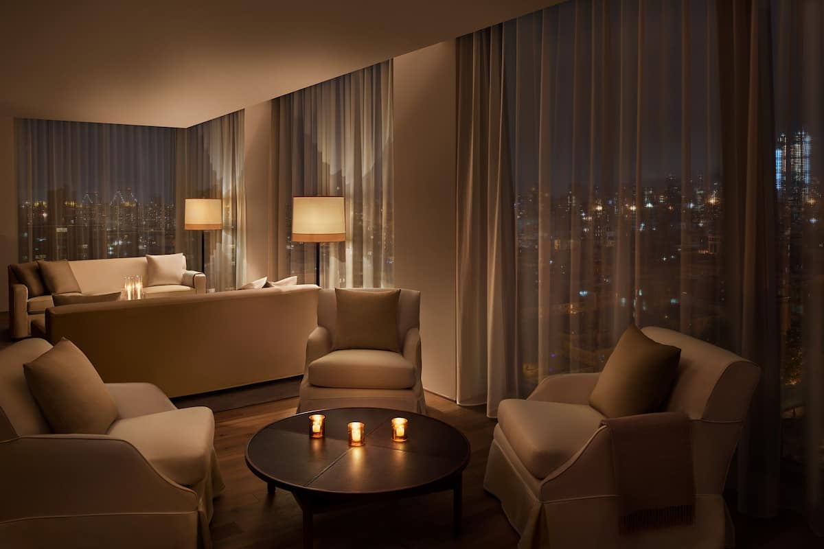 A modern hotel room with a sofa set, armchairs, a coffee table with candles, and large windows offering a city view.
