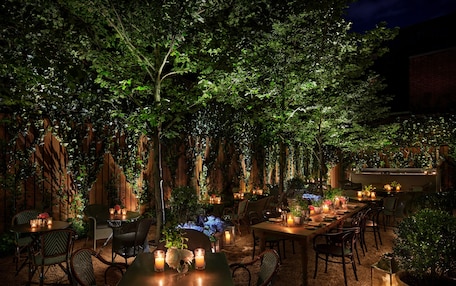 An outdoor dining area with tables and chairs, surrounded by lush greenery and wooden walls.