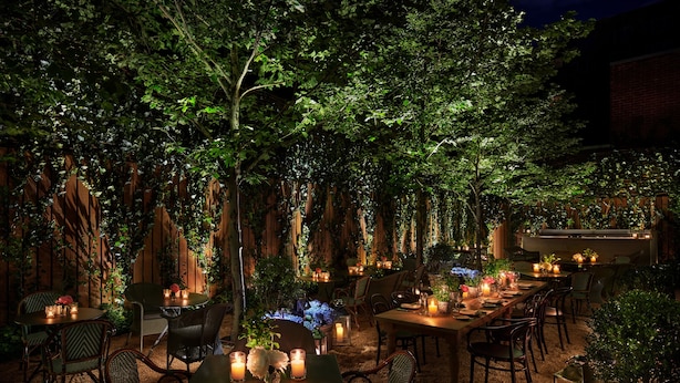 An outdoor dining area with tables and chairs, surrounded by lush greenery and wooden walls.