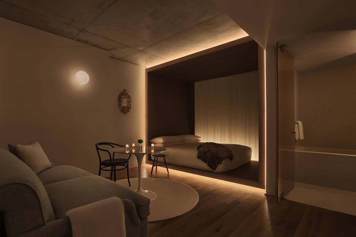A modern, minimalist bedroom with a large bed, a small table with candles, and a cozy seating area.