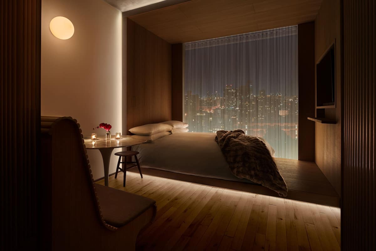 A modern bedroom with a city view, a bed, a bedside table with candles, and a chair.