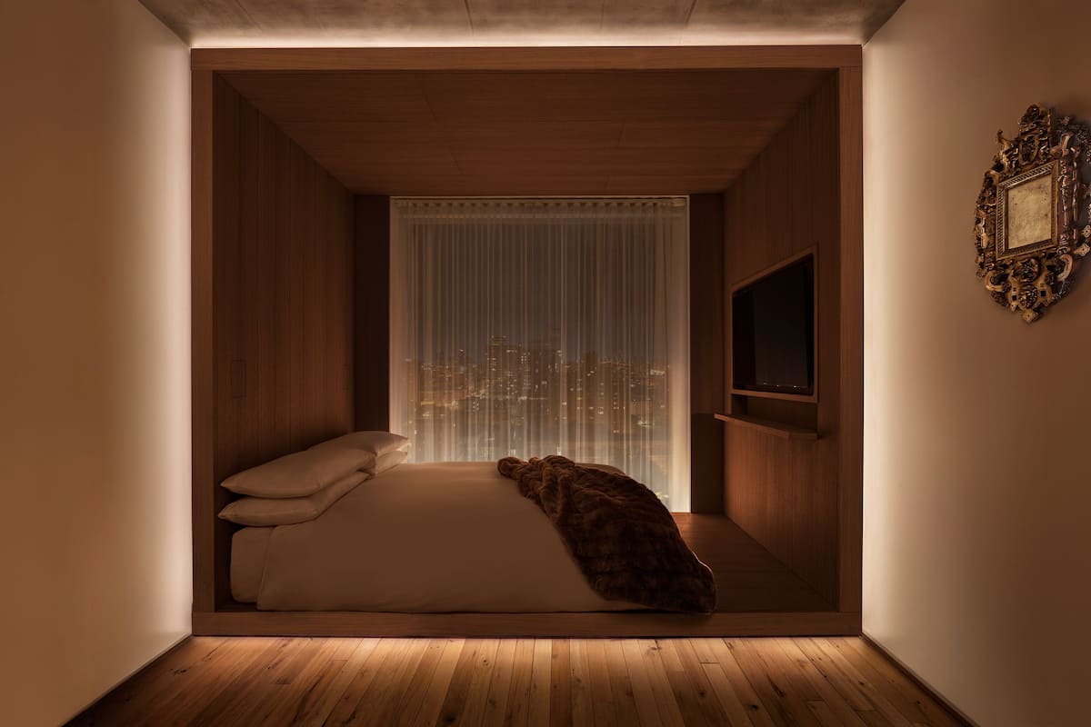 A cozy, narrow room with a bed, a window with curtains, and a television on the wall.