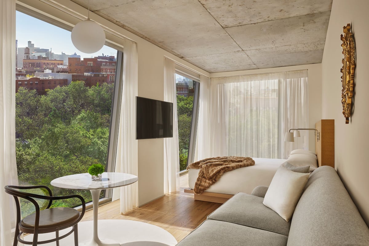 A modern hotel room with a large window, a flat-screen TV, a sofa, a small table, and a chair.