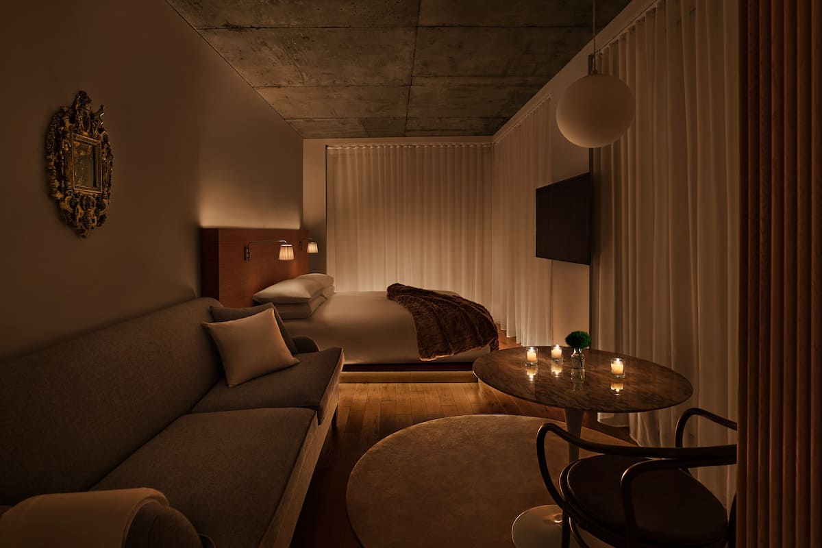 A modern hotel room with a sofa, a bed, a round table with candles, and a TV mounted on the wall.