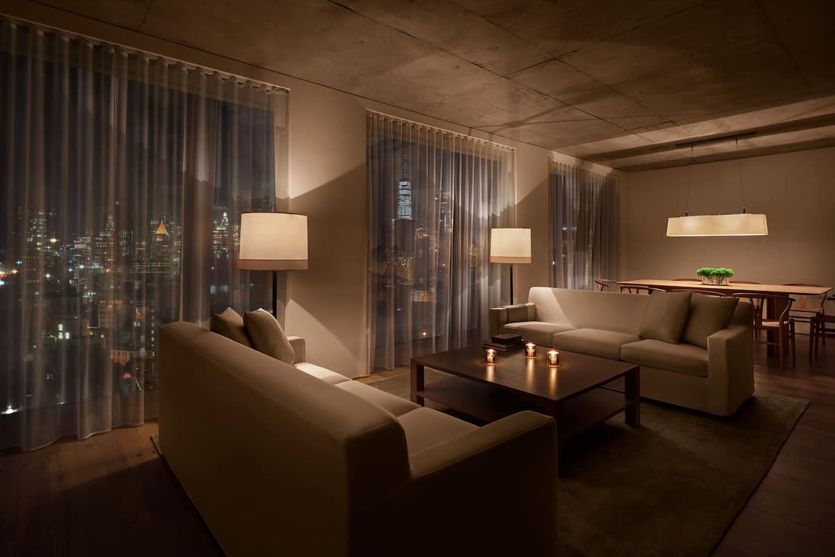A modern living room with a sofa, armchairs, a coffee table, and a dining area with a table and chairs. Large windows with sheer curtains offer a view of the cityscape at night.