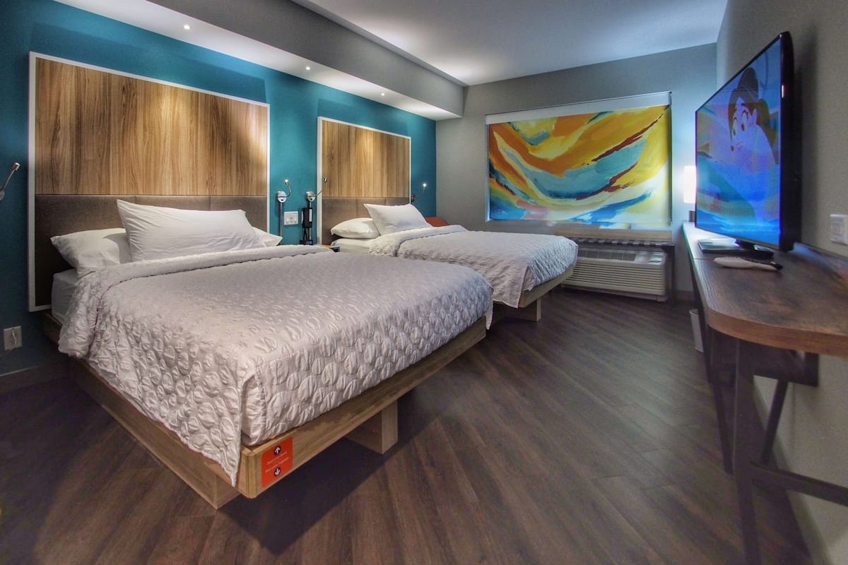 Quarto de hotel moderno com duas camas, um quadro grande e uma TV em uma escrivaninha de madeira.