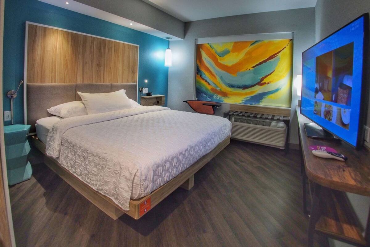 Quarto de hotel moderno com uma cama grande, televisão de tela plana, um quadro na parede e cabeceira de madeira.