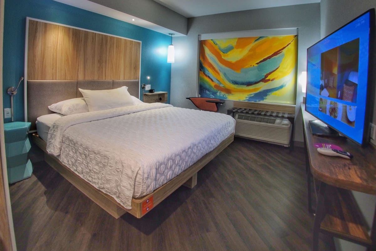 Quarto de hotel moderno com uma cama grande, televisão de tela plana, um quadro na parede e cabeceira de madeira.