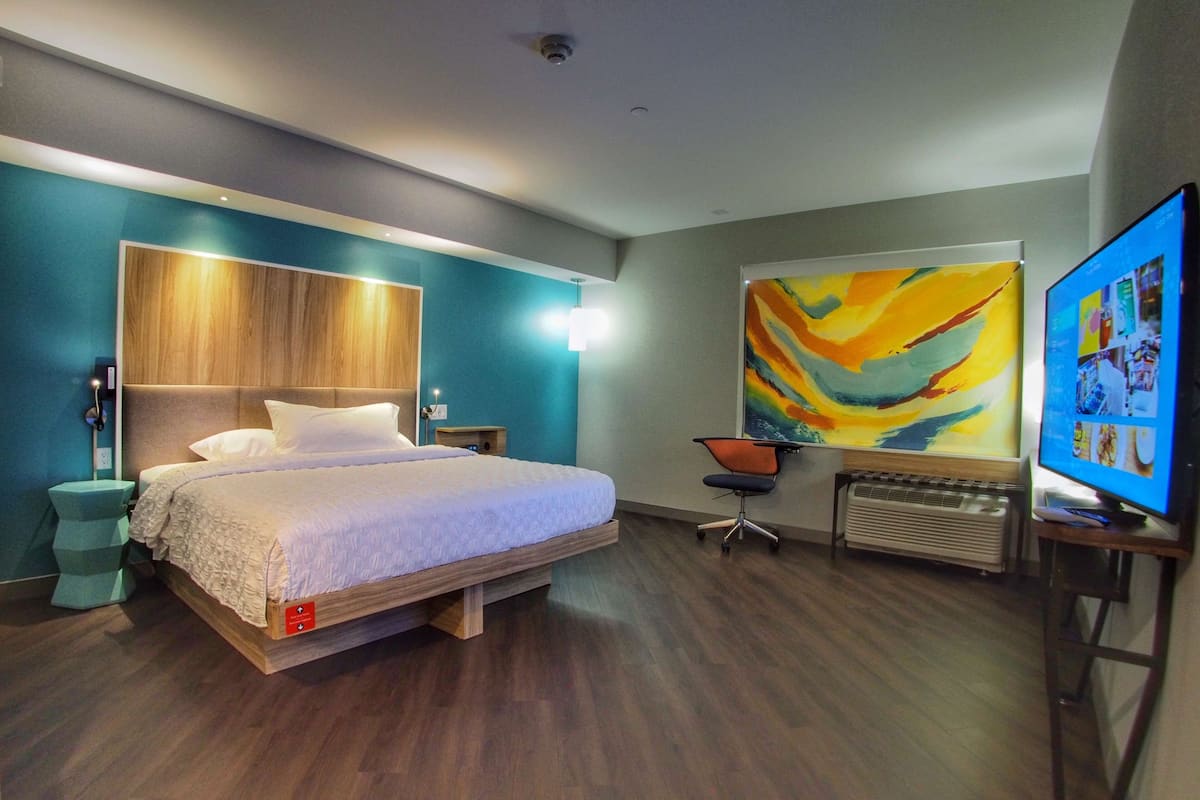 Quarto de hotel moderno com cama de madeira, uma grande pintura abstrata, uma escrivaninha com cadeira e televisão.
