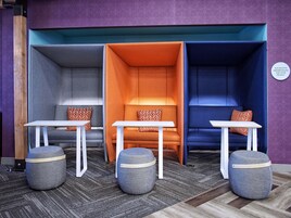 A modern lounge area with three seating pods in grey, orange, and blue, each with a small table and matching ottoman.