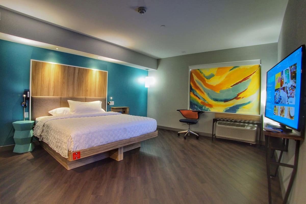 Quarto de hotel moderno com cama, escrivaninha, cadeira, pintura grande e televisão.