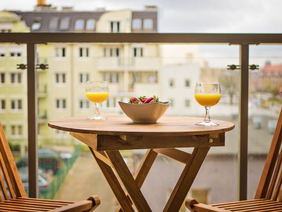 A wooden table with two glasses of orange juice and a bowl of fruit on a balcony with a city view.