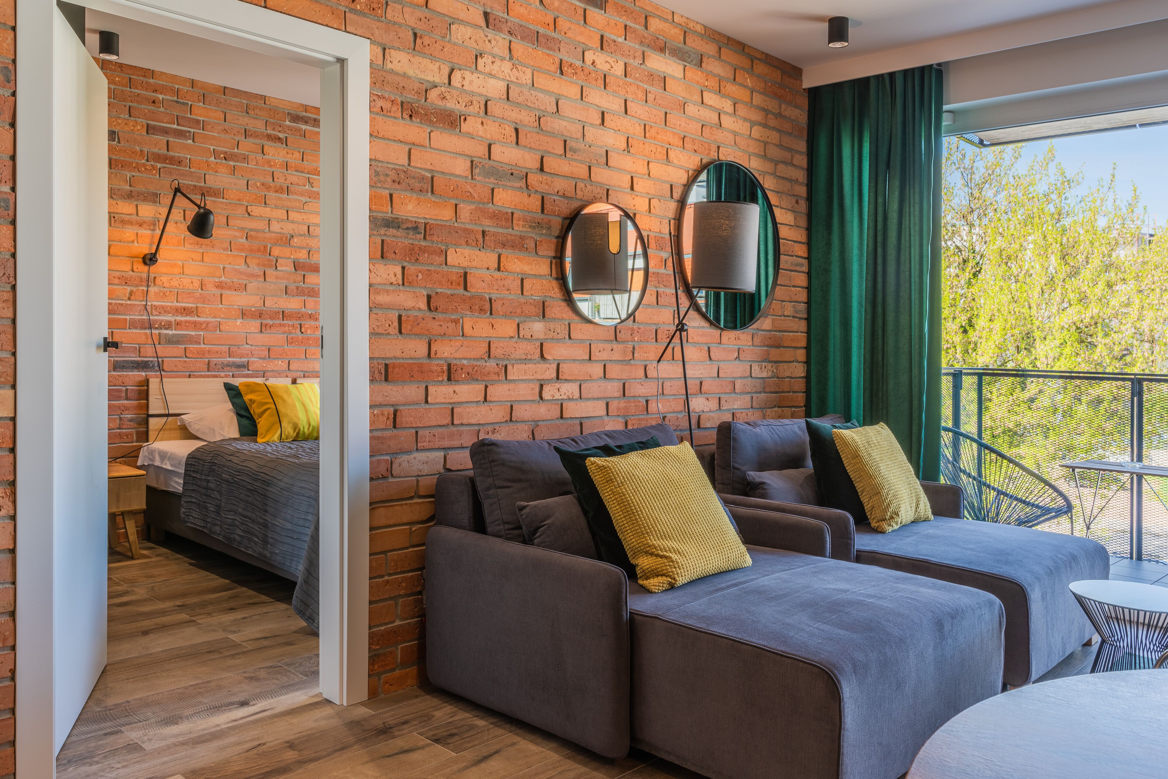A modern room with a brick wall, a sofa set, a bed, and a balcony with a view of trees.