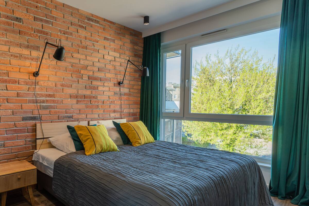 Ein Schlafzimmer mit Backsteinwand, großem Fenster mit grünen Vorhängen, einem hölzernen Nachttisch und einem Bett mit gestreifter Bettdecke und Kissen.