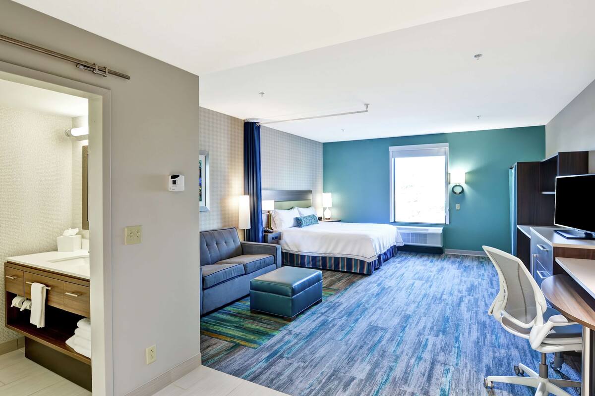 Habitación de hotel con cama, sofá, escritorio, silla, baño con lavamanos y espejo, y ventana con cortinas.