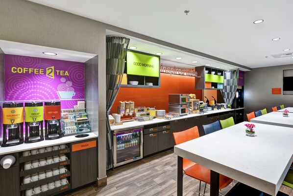 Una estación moderna para café y té, con varias máquinas de café, un refrigerador y una barra con diversos artículos.