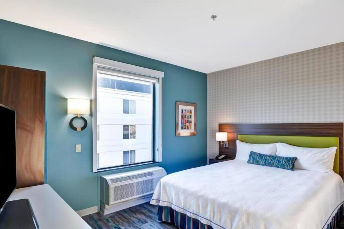 A hotel room with a bed, a desk, a window, and a wall-mounted picture.
