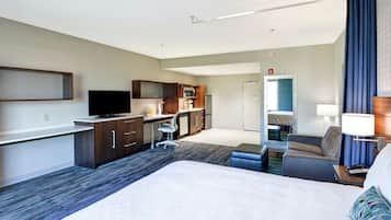 Una habitación de hotel con una cama grande, un escritorio con televisor de pantalla plana, una kitchenette y una zona de estar con sofá y sillón.