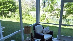 A conservatory with a view of a grassy area and trees. There is a chair with a cushion and a small table with a lamp.