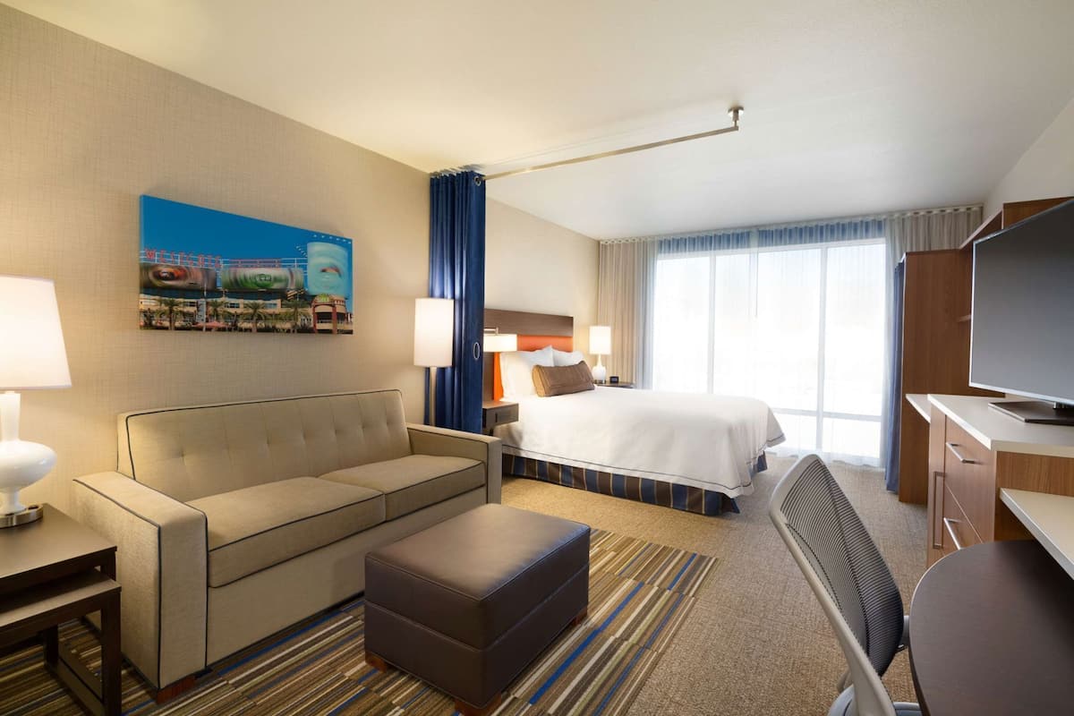Habitación de hotel con cama, sofá, reposapiés, escritorio, televisor y un cuadro en la pared.