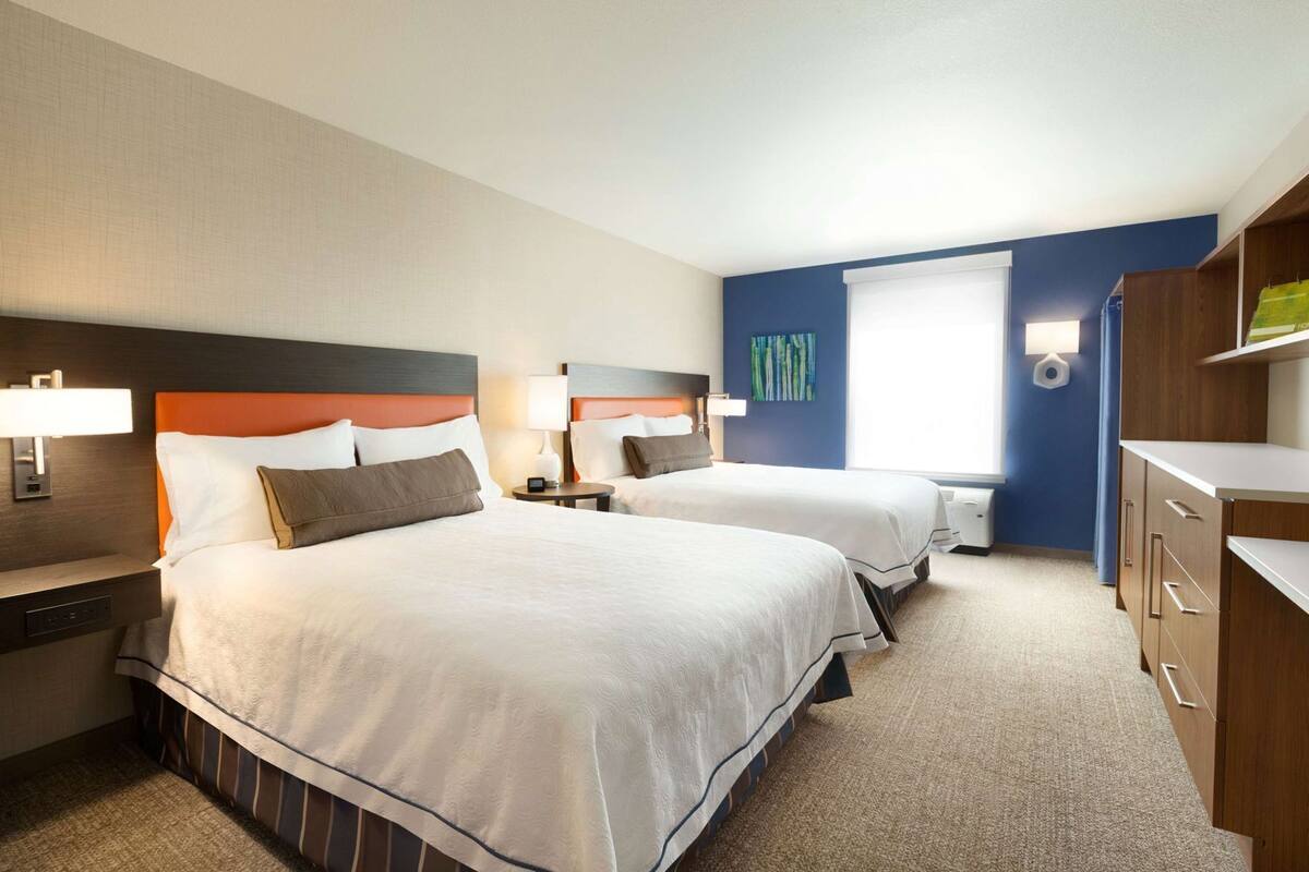 Habitación de hotel con dos camas, mesitas de noche, una lámpara, una ventana y una pared de acento azul.