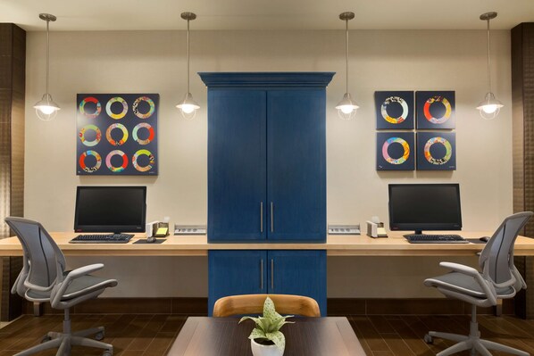 A modern office space with two desks, each with a computer and chair, a large blue cabinet, and artwork on the walls. - Home2 Suites by Hilton Glendale - Westgate (Glendale)