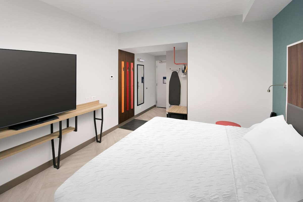 Una habitació d'hotel moderna amb un llit gran, un televisor de pantalla plana sobre un suport de fusta, un escriptori petit i una porta que dóna a un bany.