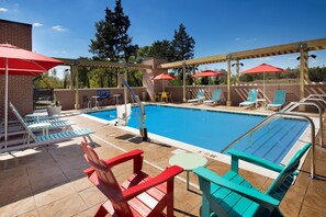 Zona de piscina con una piscina azul cristalina, sillas de descanso rojas y azules, y sombrillas rojas.