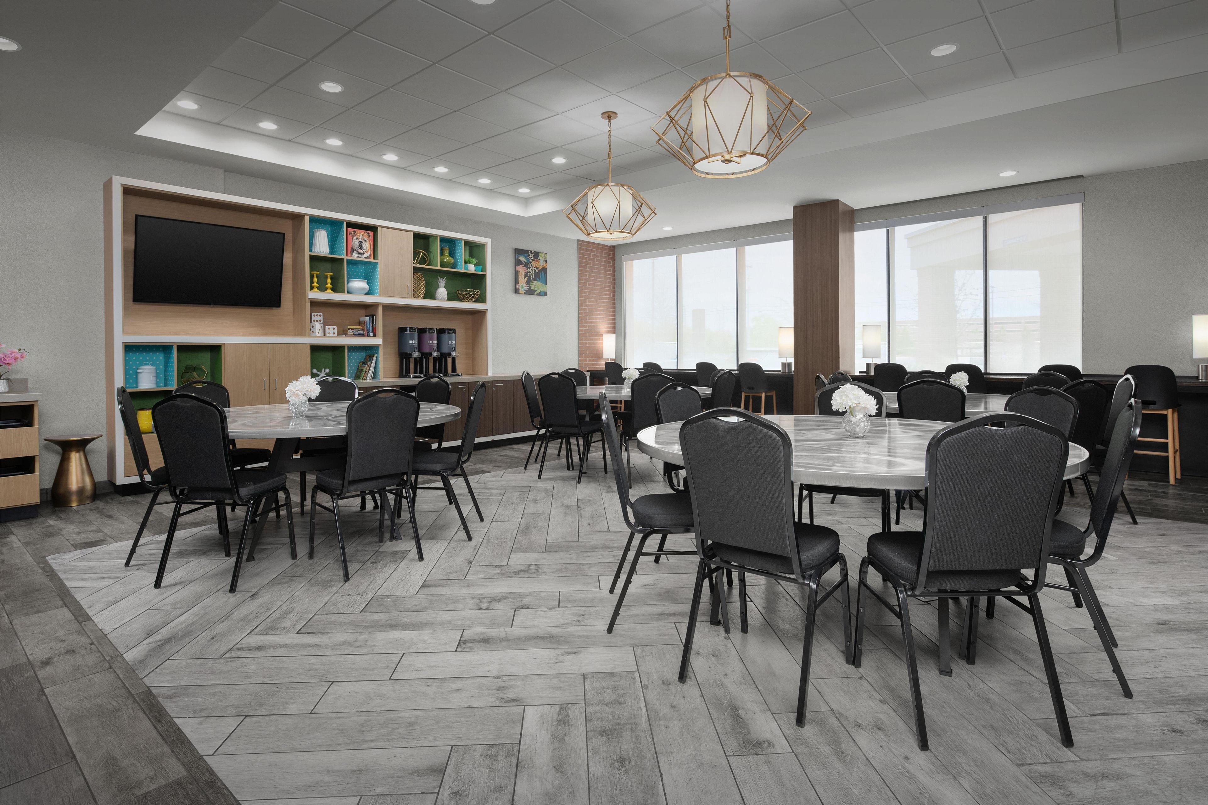 A modern conference room with multiple tables and chairs, a flat-screen TV mounted on the wall, and a large window providing natural light.