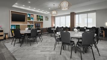 A modern conference room with multiple tables and chairs, a flat-screen TV mounted on the wall, and a large window providing natural light.