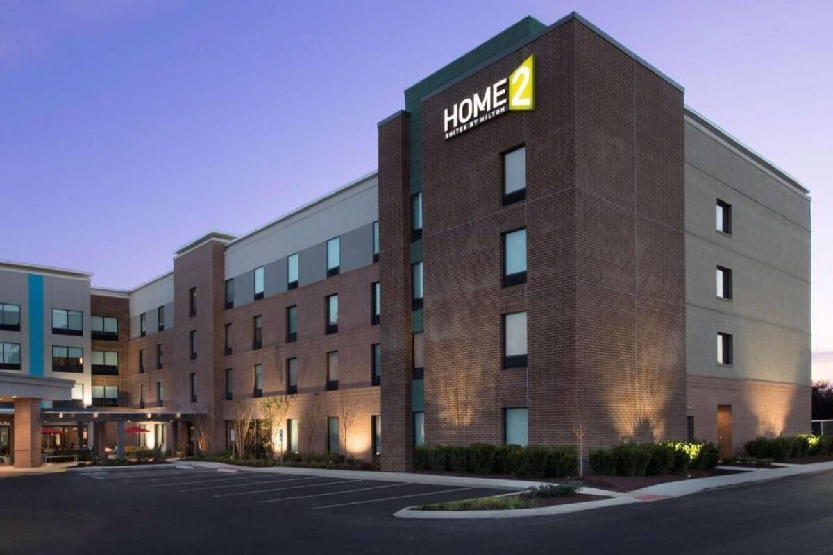 A multi-story hotel with a prominent 'HOME 2' sign, a parking lot, and a landscaped area with shrubs.