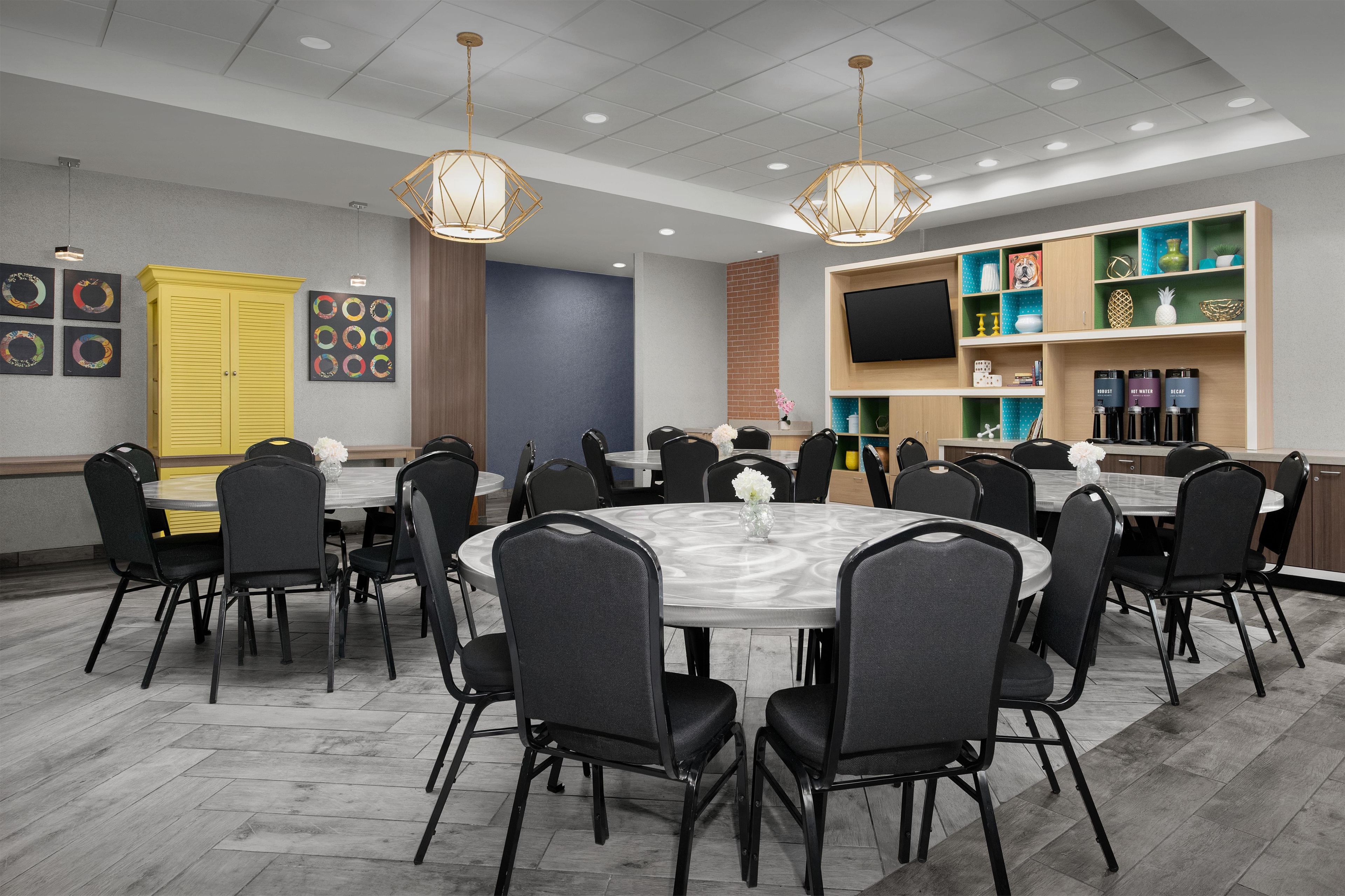 A conference room with round tables and chairs, a TV on the wall, and a shelf with various items.