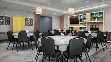 A conference room with round tables and chairs, a TV on the wall, and a shelf with various items.