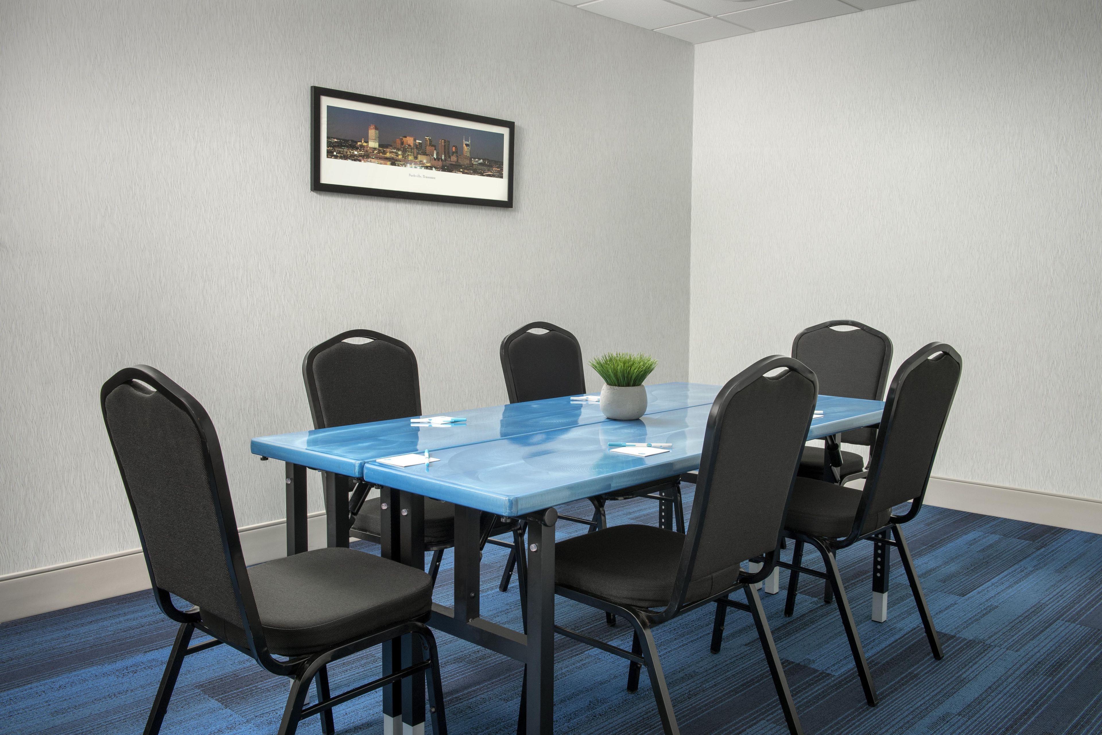 A conference room with a blue table, six chairs, a potted plant, and a framed cityscape picture on the wall.