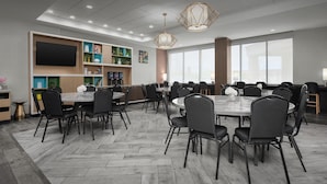 A conference room with multiple tables and chairs, a large flat-screen TV, and a decorative shelf with various items. - Home2 Suites by Hilton Murfreesboro (Murfreesboro)